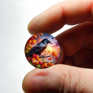 May include: A round glass cabochon with a colorful image of a black raven perched on a branch with autumn leaves. The background is a sunset sky with clouds. The text "NIX CREATIONS" appears twice on the cabochon.
