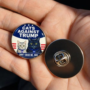 Può includere: Una spilla rotonda in metallo con un design blu, bianco e rosso con tre stelle bianche. Raffigura un gatto grigio tigrato e un gatto nero, con il testo "CATS AGAINST TRUMP" e "DON'T GRAB ME, BRO" stampato in bianco.