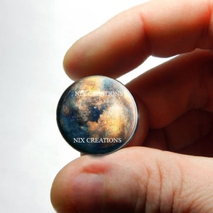 May include: A round glass cabochon with a colorful image of a planet with a blue, brown, and orange surface. The image has the text "NIX CREATIONS" printed twice on the surface.