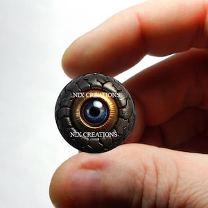 May include: A black and gold eye-shaped charm with a blue iris and the text "NIX CREATIONS" on the front and back.