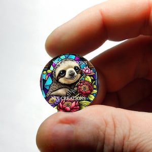 May include: A round glass cabochon with a stained glass design featuring a sloth with flowers. The sloth is brown and has a pink nose. The flowers are pink, purple, and blue. The text "NIX CREATIONS" appears twice on the cabochon.