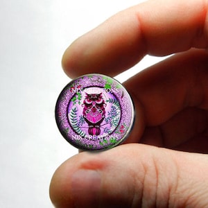 May include: A round glass cabochon with a pink owl design. The owl is surrounded by a purple and green floral pattern. The cabochon is set in a silver-toned metal setting.