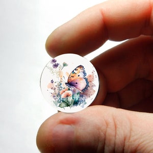 Puede incluir: Un cabujón redondo de vidrio transparente con un diseño de acuarela de una mariposa con alas naranja y rosa, rodeado de flores moradas y rosas.