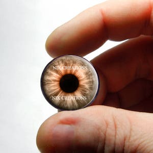 May include: A brown eye with a dark pupil, printed on a round glass cabochon. The cabochon is held in a hand, and the text "NIX CREATIONS" is printed on the glass.