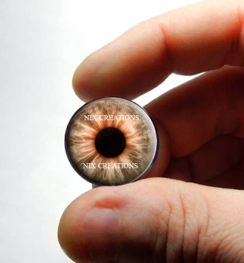 May include: A brown eye with a dark pupil, printed on a round glass cabochon. The cabochon is held in a hand, and the text "NIX CREATIONS" is printed on the glass.