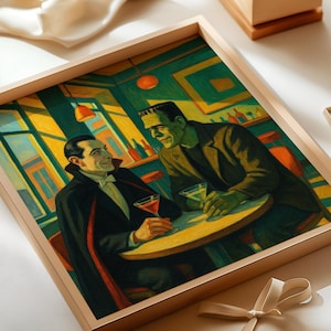 May include: Framed artwork depicting Dracula and Frankenstein at a bar, each holding a cocktail. The painting features a warm color palette with green, yellow, and red accents. The artwork is in a light-colored wooden frame.