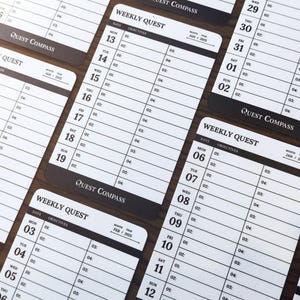 May include: Several white and black weekly planner cards with the text "Weekly Quest" and "Quest Compass". Each card has dates and space for objectives. The cards are arranged on a dark brown surface.
