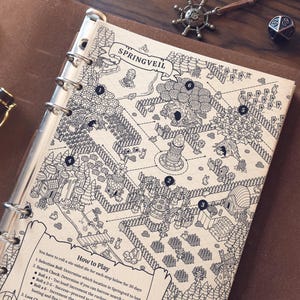May include: A detailed black and white map of Springveil, a fantasy game, is displayed in a binder. The map features numbered locations, trees, buildings, and a banner with the game's name. A pen, dice, and a compass charm are also visible.