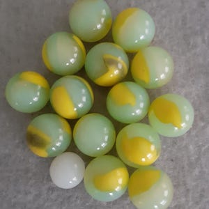 May include: A selection of glass marbles in pale green and yellow hues. The marbles have a swirled design, with some featuring a solid yellow pattern. One white marble is also present.