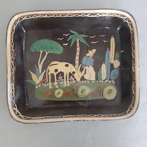 Op de afbeelding: Rechthoekige, handbeschilderde decoratieve schaal met een zwarte achtergrond en een crèmekleurige rand. De schaal toont een pastorale scène met een boom, palmboom en figuren. De scène omvat een koe, een persoon en cactussen.