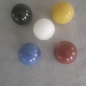 Puede incluir: Cinco canicas de colores: negro, blanco, amarillo, azul y rojo. Cada canica es una esfera lisa y brillante. Las canicas están dispuestas sobre una superficie gris claro.