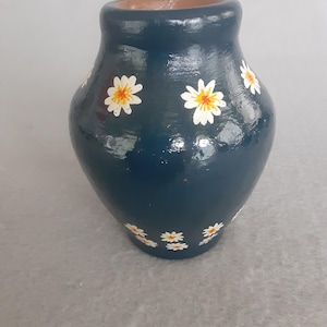 May include: A dark teal vase with a glossy finish, adorned with hand-painted white daisies with yellow centers. The vase has a rounded body and a slightly flared rim, suitable for displaying flowers.