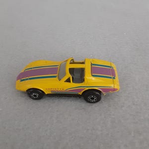 Puede incluir: Un coche de juguete Corvette amarillo con rayas moradas y turquesas. El coche tiene ruedas negras y la palabra "CORVETTE" impresa en el lateral. Es una réplica en miniatura de un coche deportivo clásico.