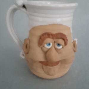 May include: A white ceramic mug with a sculpted face design. The face is tan with brown hair, a mustache, and blue eyes. The handle has a small white decorative element. A unique and whimsical coffee mug.