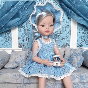 May include: A blue crocheted dress and bonnet set for a doll. The dress has a white lace trim and a blue ribbon. The bonnet has a white lace trim and a blue ribbon. The doll is sitting on a blue couch with a snow globe in her hands.