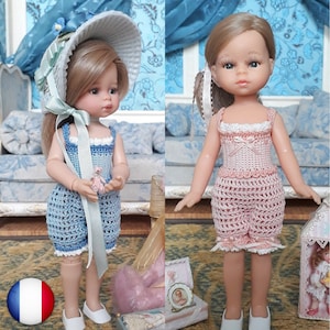 May include: Two dolls wearing crocheted dresses. The doll on the left is wearing a light blue dress with a white crocheted hat. The doll on the right is wearing a pink dress. Both dolls have blonde hair and blue eyes.