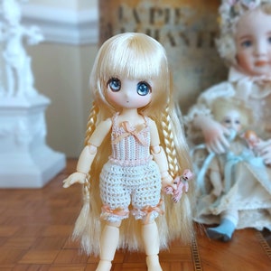 Op de afbeelding: Een kleine pop met lang blond haar die een gehaakt roze en wit jurkje draagt en een klein knuffeldier vasthoudt. De pop heeft blauwe ogen en een roze strik in haar haar.