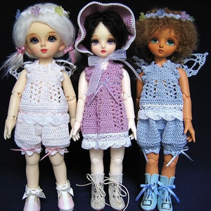 May include: Three dolls wearing crocheted outfits. The doll on the left is wearing a white crocheted dress and shorts with pink accents. The doll in the middle is wearing a purple crocheted dress with white lace trim. The doll on the right is wearing a blue crocheted dress and shorts with white lace trim.