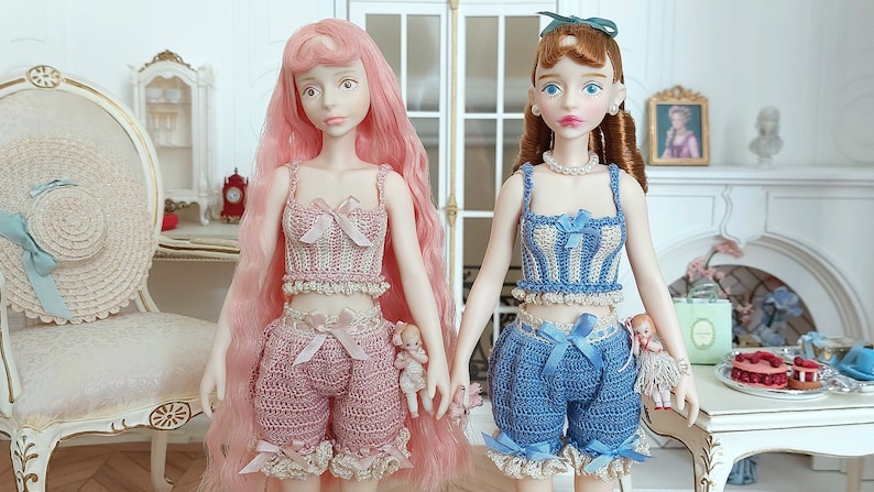 May include: Two fashion dolls are holding hands, each wearing a crocheted outfit. One doll has pink hair and a pink top and shorts. The other doll has brown hair and a blue and white top and blue shorts. Both dolls have small dolls attached to their outfits.