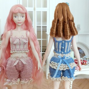 May include: Two dolls wearing handmade crochet outfits. One doll has long pink hair and a pink outfit with a bow. The other doll has brown hair and a blue and white outfit with a bow. Both dolls are standing in a room with a table.