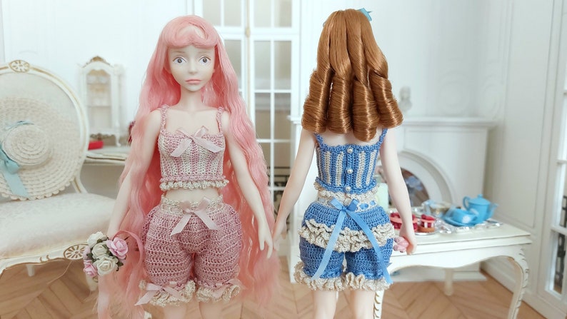 May include: Two dolls wearing handmade crochet outfits. One doll has long pink hair and a pink outfit with a bow. The other doll has brown hair and a blue and white outfit with a bow. Both dolls are standing in a room with a table.