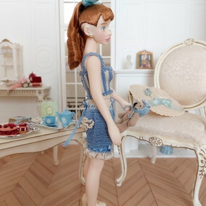 May include: A doll with auburn hair in a blue and cream crochet outfit, platform shoes, and a matching bow. The doll holds a small teddy bear. A straw hat with a blue ribbon sits on a chair. The scene suggests a dollhouse or miniature setting.
