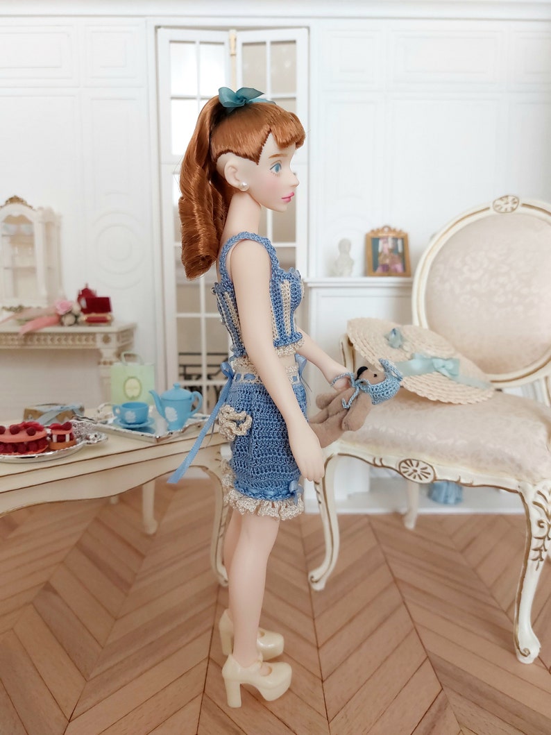 May include: A doll with auburn hair in a blue and cream crochet outfit, platform shoes, and a matching bow. The doll holds a small teddy bear. A straw hat with a blue ribbon sits on a chair. The scene suggests a dollhouse or miniature setting.