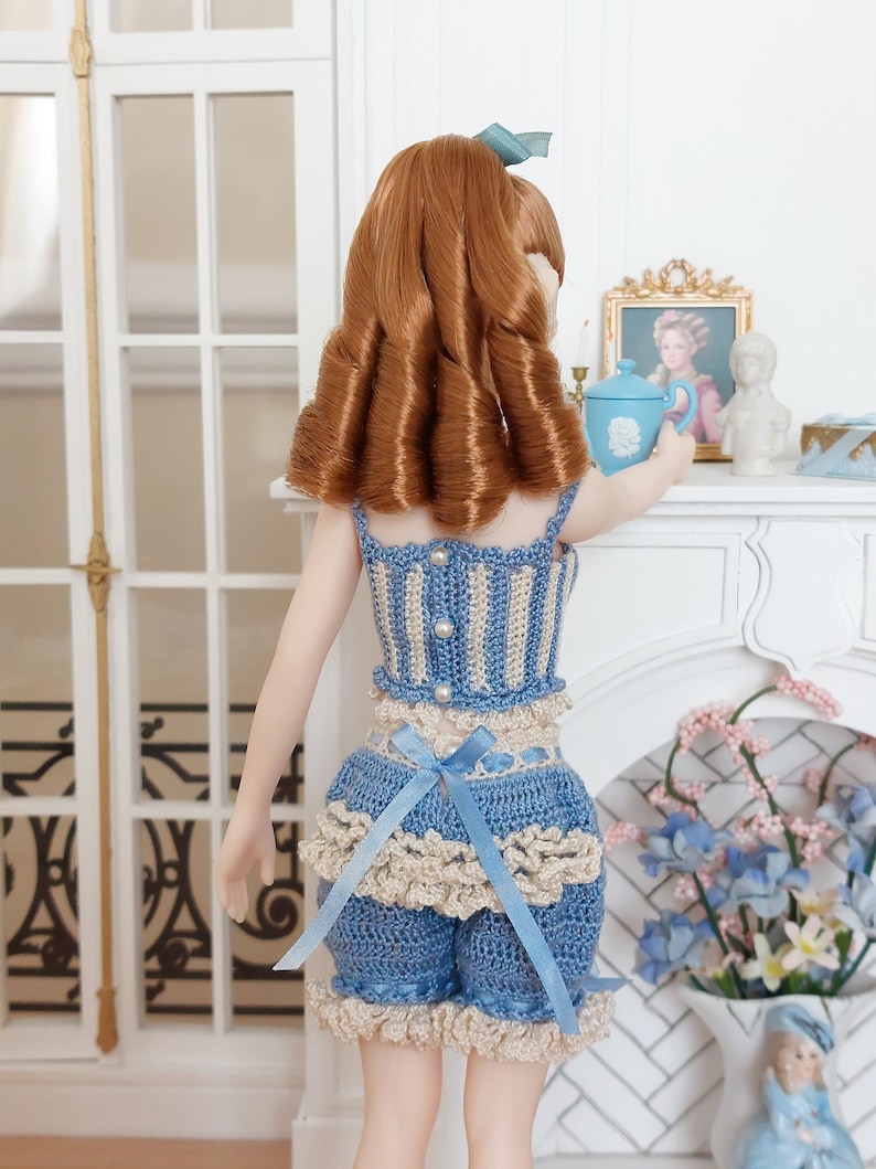 May include: A doll wearing a blue and cream crochet outfit with a matching bow. The outfit includes a top and shorts, both with a ruffled trim. The doll has auburn hair and is holding a blue teacup. The scene is set in a miniature room with a fireplace.
