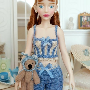 May include: A doll wearing a blue and cream crochet top and shorts set with blue bows. The doll has red hair and is holding a small teddy bear wearing a blue bonnet. A matching hat is on a nearby chair.