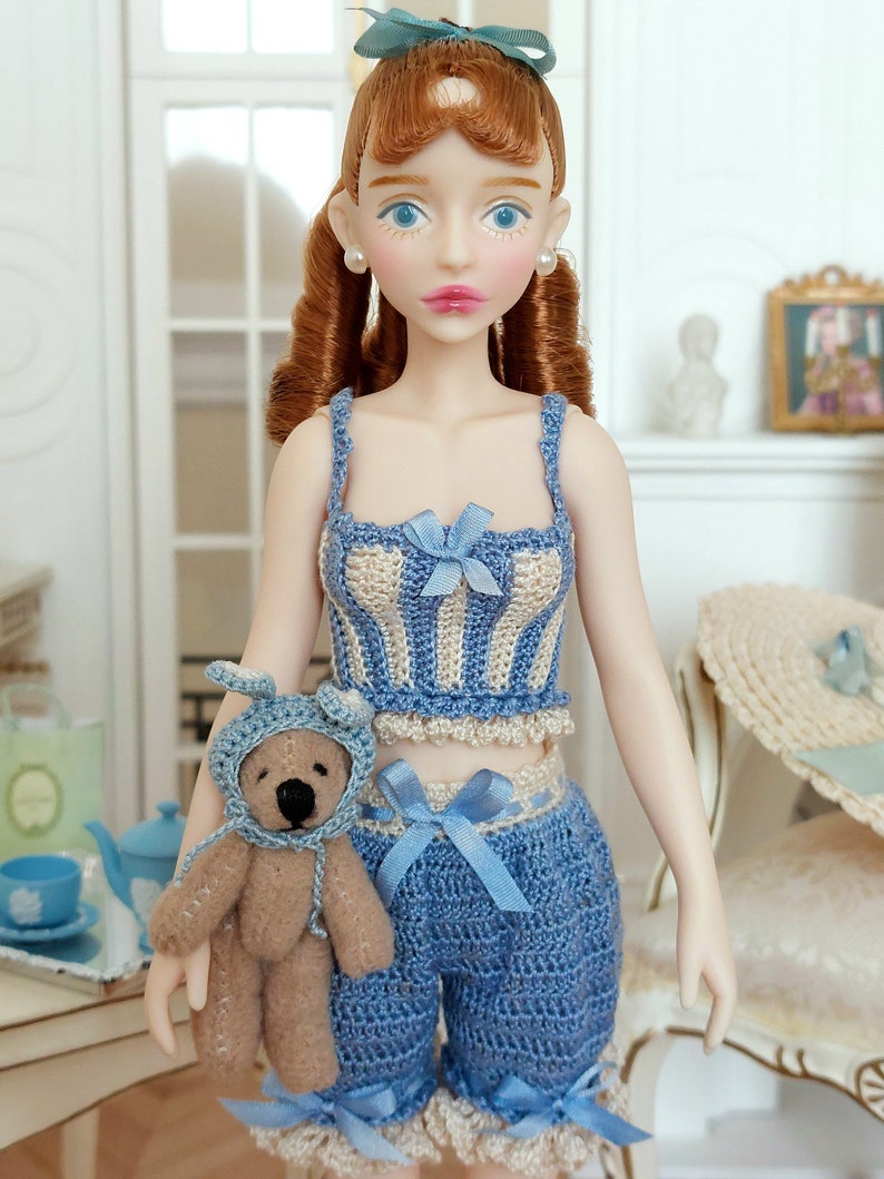 May include: A doll wearing a blue and cream crochet top and shorts set with blue bows. The doll has red hair and is holding a small teddy bear wearing a blue bonnet. A matching hat is on a nearby chair.
