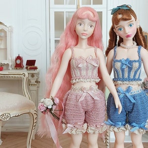 May include: Two dolls wearing matching crochet outfits. One doll has pink hair and a pink outfit with a bow. The other doll has brown hair and a blue outfit with a bow. A straw hat with a blue ribbon sits on a chair.