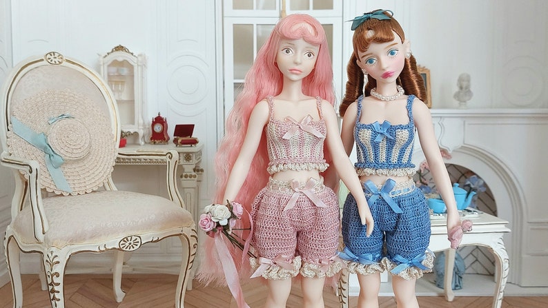May include: Two dolls wearing matching crochet outfits. One doll has pink hair and a pink outfit with a bow. The other doll has brown hair and a blue outfit with a bow. A straw hat with a blue ribbon sits on a chair.