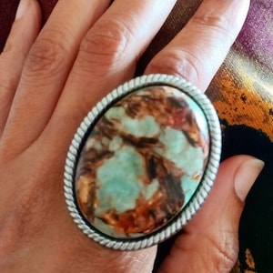 May include: A silver ring with a large oval-shaped stone. The stone is a mix of brown, green, and white colors, with a marbled pattern.