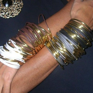Puede incluir: Una pila de brazaletes dorados y blancos con una pila de brazaletes dorados y grises en el brazo de una persona.