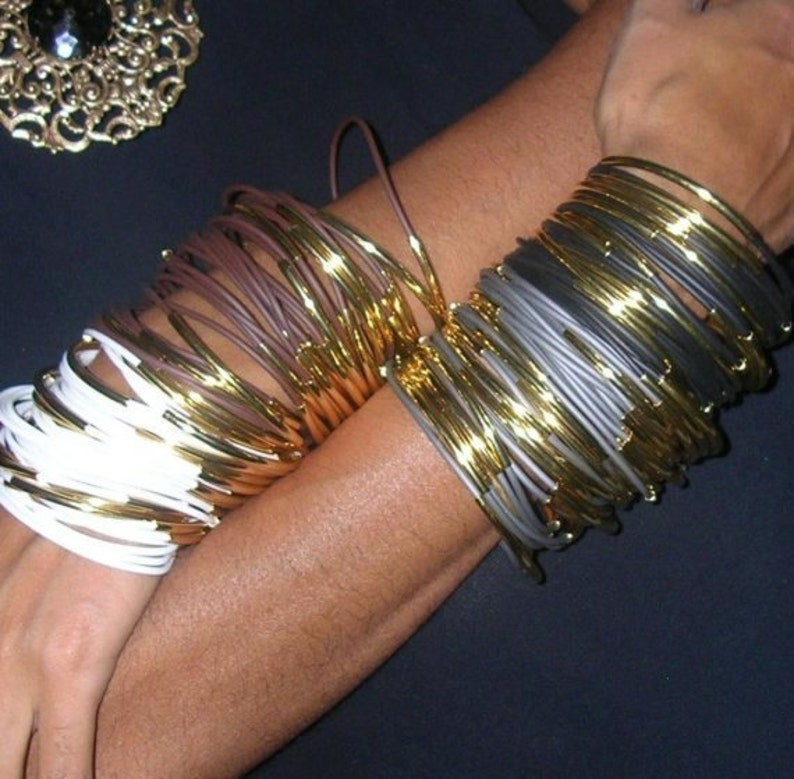 Puede incluir: Una pila de brazaletes dorados y blancos con una pila de brazaletes dorados y grises en el brazo de una persona.