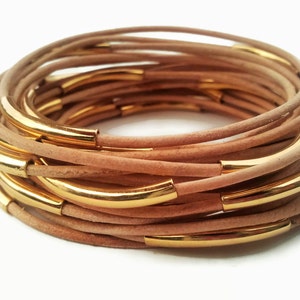 May include: A stack of beige leather bracelets with gold metal accents. The bracelets are layered on top of each other, creating a unique and stylish look.