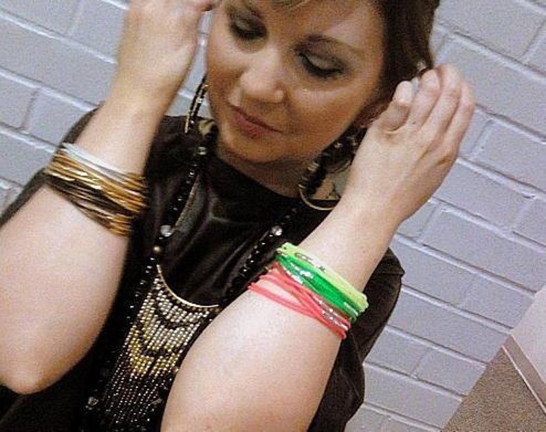 Puede incluir: Una mujer con un top negro, un collar de cuentas y varios brazaletes en ambas mu&ntilde;ecas. Los brazaletes son una mezcla de colores dorados, plateados y de ne&oacute;n.