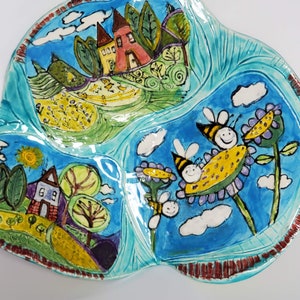 May include: A whimsical, hand-painted ceramic serving dish with three sections. The dish features a vibrant turquoise background with colorful illustrations of houses, trees, bees, and flowers. The rim has a textured, reddish-brown border, adding a rustic touch to this decorative piece.