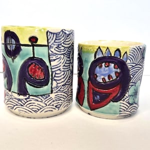 May include: Two ceramic mugs with colorful, abstract designs. The mugs are decorated with blue, red, green, and purple paint. The designs feature geometric shapes and floral motifs.