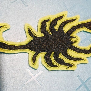 May include: A black and gold scorpion patch with a yellow outline. The scorpion design is embroidered with a textured appearance. The patch is likely for clothing or accessories.