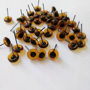 May include: A group of amber-colored plastic safety eyes with black pupils and metal posts. The eyes are scattered on a white surface.
