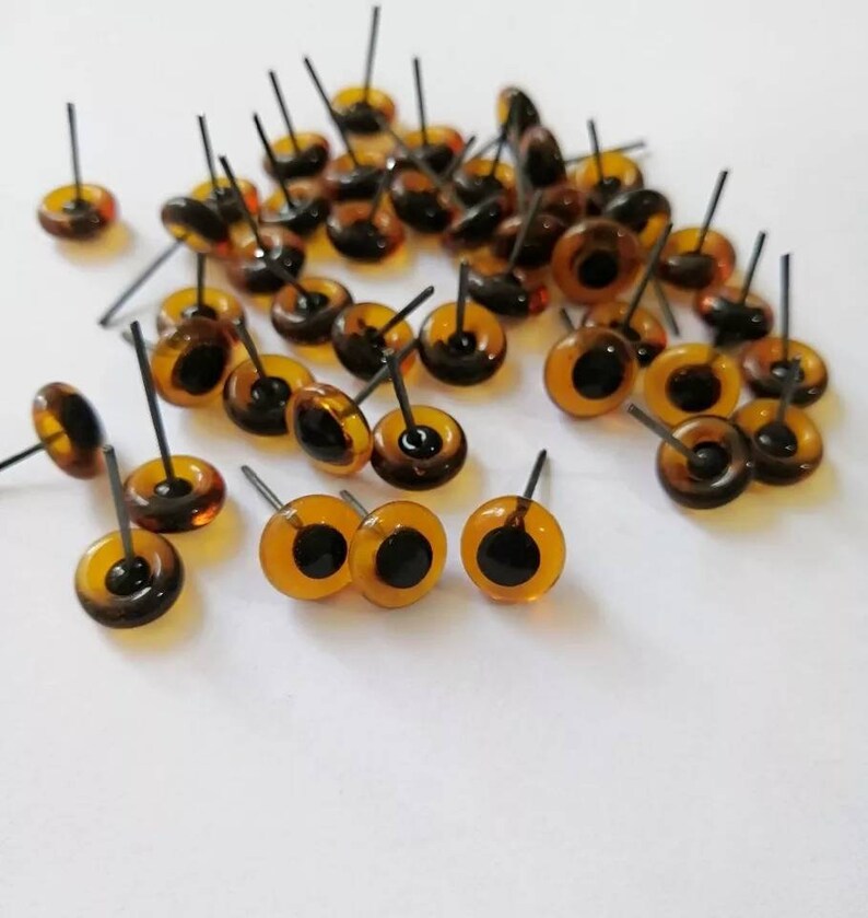 May include: A group of amber-colored plastic safety eyes with black pupils and metal posts. The eyes are scattered on a white surface.