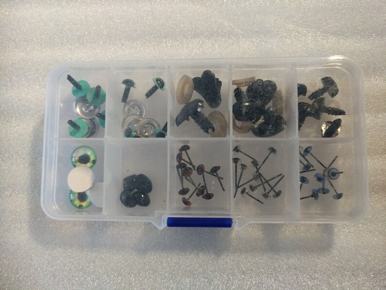 May include: A clear plastic storage box with multiple compartments filled with various sizes and colors of plastic safety eyes for crafts. The box has a blue handle.