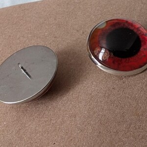 May include: Two round, red and black eye-shaped cabochons with a silver-colored metal rim. One is face up, the other is face down, showing the metal backing with a loop. The cabochons have a glossy finish.