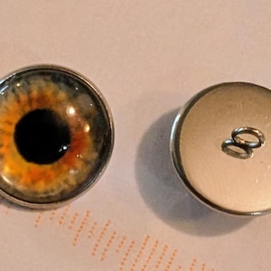 May include: Two round buttons featuring an eye design. One button displays a detailed eye with orange, yellow, and black hues, set in a silver-coloured metal. The other button is plain silver with two loops for sewing.