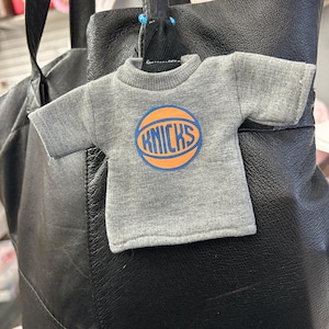 May include: A miniature grey t-shirt ornament featuring a basketball graphic with the text KNICKS in blue and orange. The small garment has short sleeves and a ribbed crew neckline, attached to a black leather bag by a small blue plastic loop.