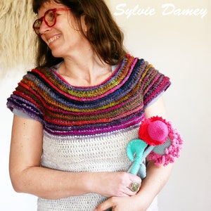 Puede incluir: Una mujer lleva un top de crochet multicolor con un sombrero de crochet blanco y pantalones vaqueros azules. El top tiene un amplio escote de colores y mangas cortas. La mujer sostiene dos flores de crochet en sus manos.