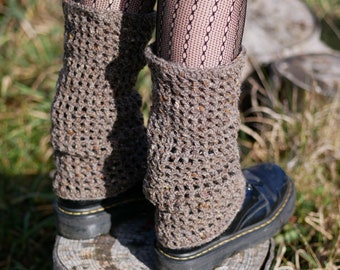 May include: A pair of brown crochet leg warmers with a textured, open weave design. The leg warmers are worn over black boots and fishnet stockings. The leg warmers are a neutral color and appear to be handmade.
