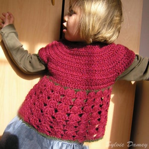 Peut inclure: Un jeune enfant portant un pull en crochet rose avec une bordure verte. Le pull a un design unique avec un bas festonné.