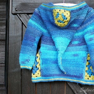 Puede incluir: Un cárdigan con capucha de color azul y verde tejido a crochet con un diseño de flor amarilla y verde en la espalda. El cárdigan está colgado en una puerta de madera.